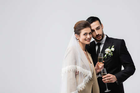 Smiling bride in veil holding glass of champagne near arabian boyfriend isolated on grayの写真素材