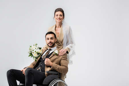 elegant woman in veil, with wedding bouquet, looking at camera near handicapped arabian fiance isolated on grayの写真素材