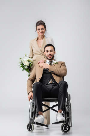 happy, disabled arabian man and elegant bride with wedding bouquet looking at camera on grayの写真素材