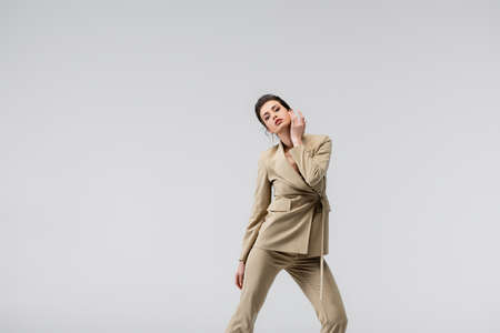 brunette woman in stylish pantsuit touching face while posing isolated on grayの写真素材