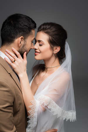 joyful woman in veil touching face of arabian bridegroom isolated on grayの写真素材