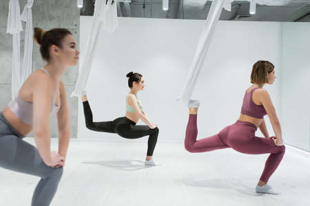 sportive women stretching with fly yoga hammocks in fitness center, blurred foregroundの写真素材
