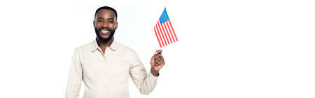 joyful african american man holding small flag of usa and looking at camera isolated on white,...