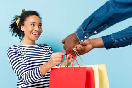 cheerful african american wife taking shopping bags from husband on blueの写真素材