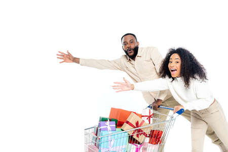 excited african american couple pointing with hands near shopping cart with gift boxes isolated on whiteの写真素材