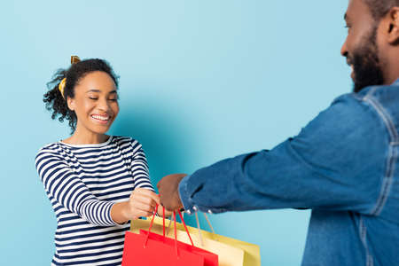 happy african american woman taking shopping bags from husband on blueの写真素材