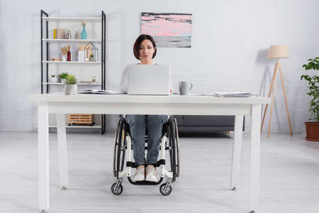 Brunette teleworker in wheelchair using laptop near notebook and papers at home