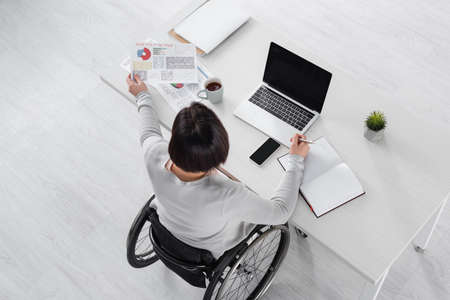 Overhead view of woman in wheelchair working with documents, devices and coffee on tableの写真素材
