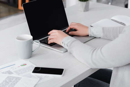 Cropped view of woman using laptop near cup, smartphone with blank screen and papers on blurred...