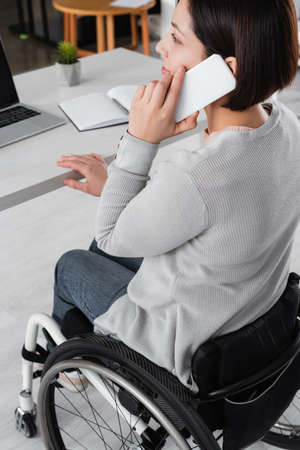 Disabled freelancer talking on smartphone near laptop and notebook on blurred background