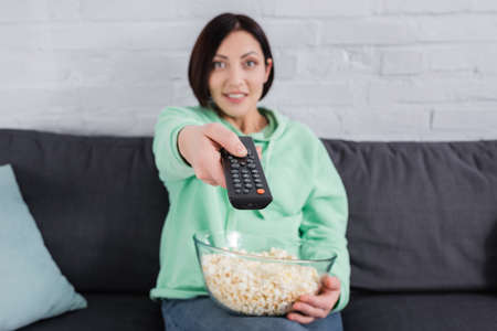 Remote controller in hand of smiling woman with popcorn on couch on blurred background