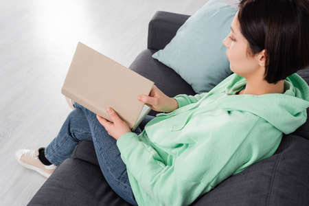 Brunette woman holding book while sitting on couch at homeの写真素材