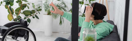 Brunette woman using virtual reality headset near wheelchair at home, banner