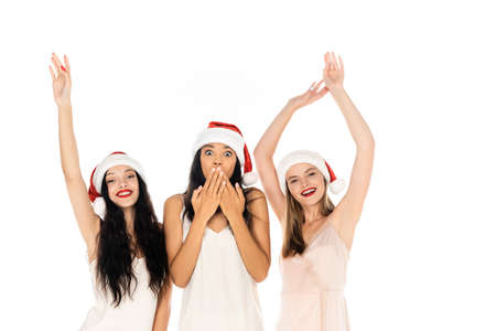 young and cheerful interracial women in santa hats and dresses standing isolated on whiteの写真素材