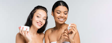 brunette multiethnic women with bare shoulders smiling while holding containers with cosmetic cream isolated on gray, bannerの写真素材