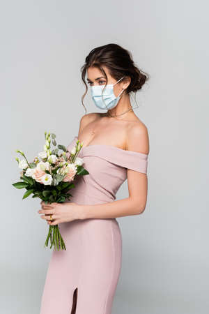 young bride in pink dress and medical mask posing with wedding bouquet isolated on grayの写真素材