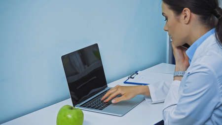 stomatologist typing on laptop and talking on smartphone near fresh apple and clipboardの写真素材
