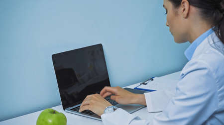 dentist typing on laptop with blank screen near fresh apple and clipboard