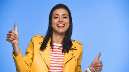 Positive woman in yellow jacket showing thumbs up gesture isolated on blueの写真素材