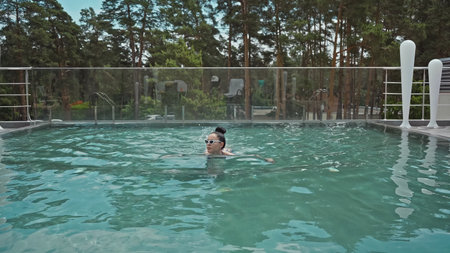 brunette young woman in sunglasses swimming in outdoor pool