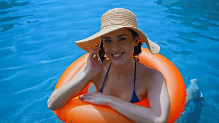 happy woman swimming in pool with inflatable ring and talking on smartphone