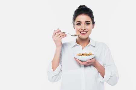 Smiling brunette woman holding spoon and bowl of cereals isolated on whiteの写真素材