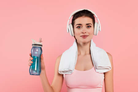 Brunette sportswoman in headphones and towel holding sports bottle isolated on pinkの写真素材
