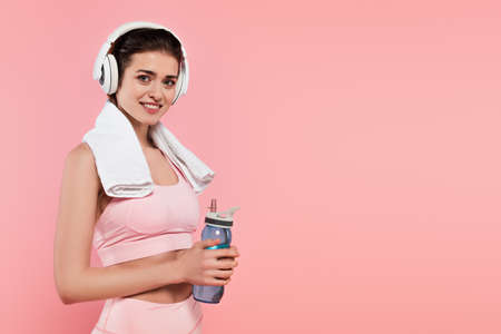 Sportswoman smiling at camera while listening music in headphones and holding sports bottle isolated on pinkの写真素材
