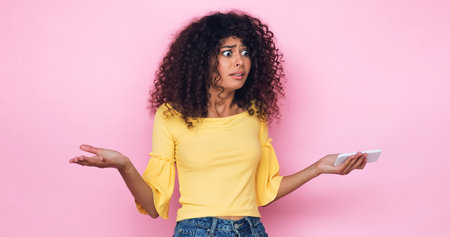 confused young woman showing shrug gesture while holding smartphone isolated on pinkの写真素材