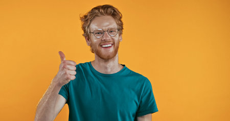 cheerful redhead man in glasses showing thumb up isolated on yellowの写真素材