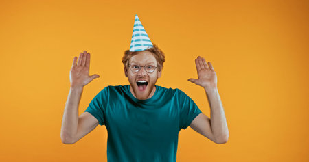amazed redhead man in party cap gesturing isolated on yellowの写真素材