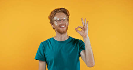 positive redhead man in glasses showing okay sign isolated on yellowの写真素材