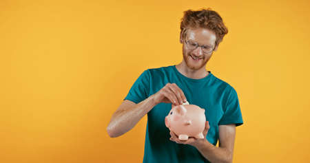 happy redhead man putting coin in piggy bank isolated on yellowの写真素材