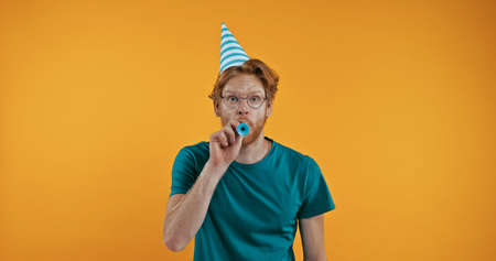 redhead man in party cap blowing in party horn isolated on yellowの写真素材