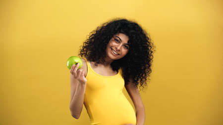 Pregnant hispanic woman with apple smiling at camera isolated on yellowの写真素材