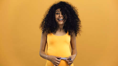 Cheerful pregnant hispanic woman holding tape measure isolated on yellowの写真素材