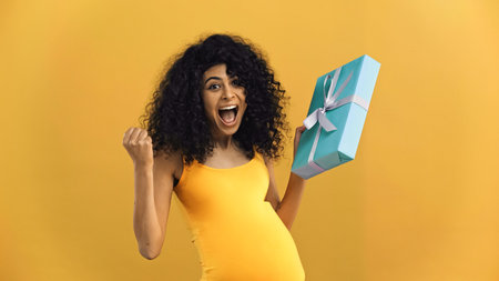 Excited pregnant hispanic woman holding present isolated on yellowの写真素材