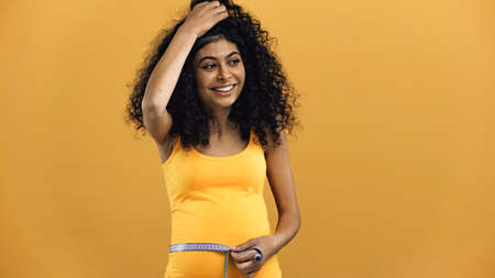 Happy pregnant hispanic woman holding tape measure isolated on yellowの写真素材