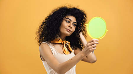 Young hispanic woman looking at mirror on blurred foreground isolated on yellowの写真素材