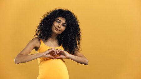 Pregnant hispanic woman showing love sign isolated on yellowの写真素材