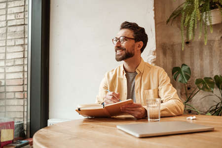 cheerful bearded freelancer in eyeglasses holding notebook and looking awayの写真素材
