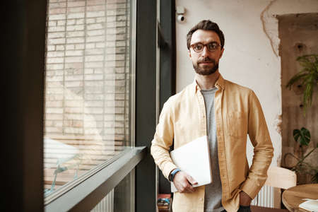 bearded freelancer holding laptop while standing with hand in pocket near windowの写真素材