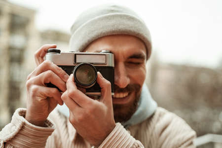 bearded photographer in beanie hat smiling while taking photo on retro cameraの写真素材