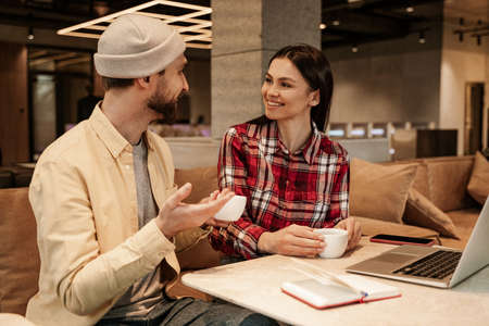 cheerful woman looking at man in beanie hat with cup of coffeeの写真素材
