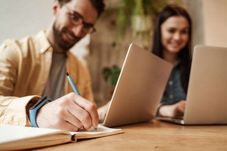 blurred freelancer in eyeglasses writing in notebook near woman using laptop in cafeの写真素材