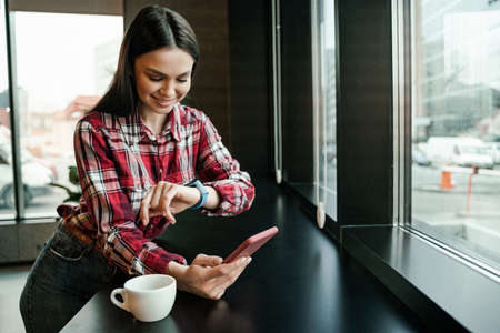 happy woman looking at smart watch while holding mobile phone near cup on tableの写真素材
