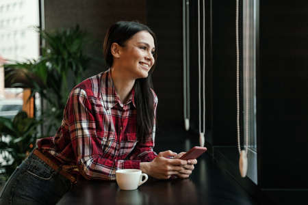 happy brunette woman leaning on table and holding smartphone