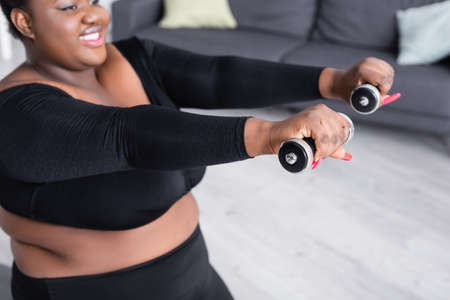 blurred african american plus size woman in sportswear exercising with dumbbells in living roomの写真素材
