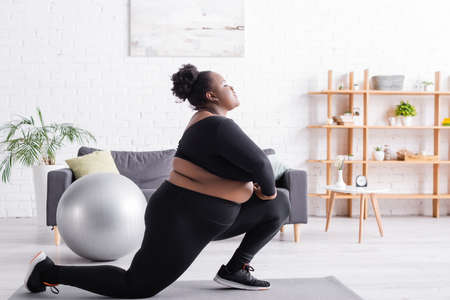 side view of african american plus size woman in sportswear doing lunges in living room