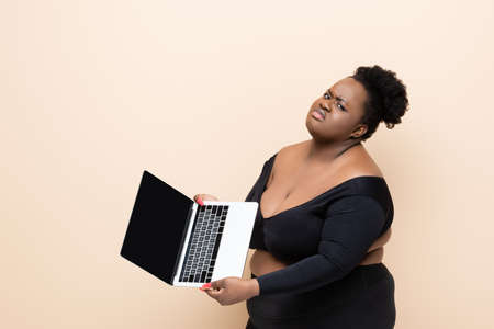 displeased african american plus size woman holding laptop isolated on beigeの写真素材